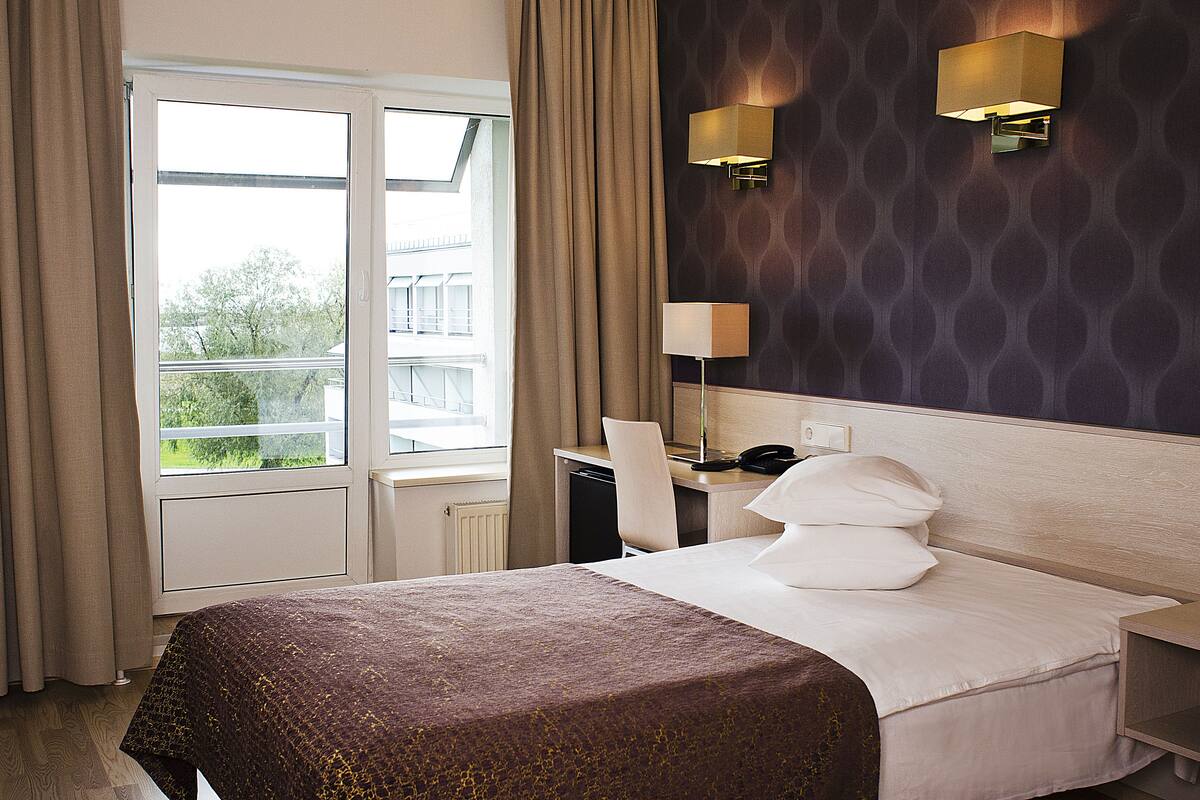 A hotel room with a bed, a desk, a chair, a lamp, and a window with curtains.