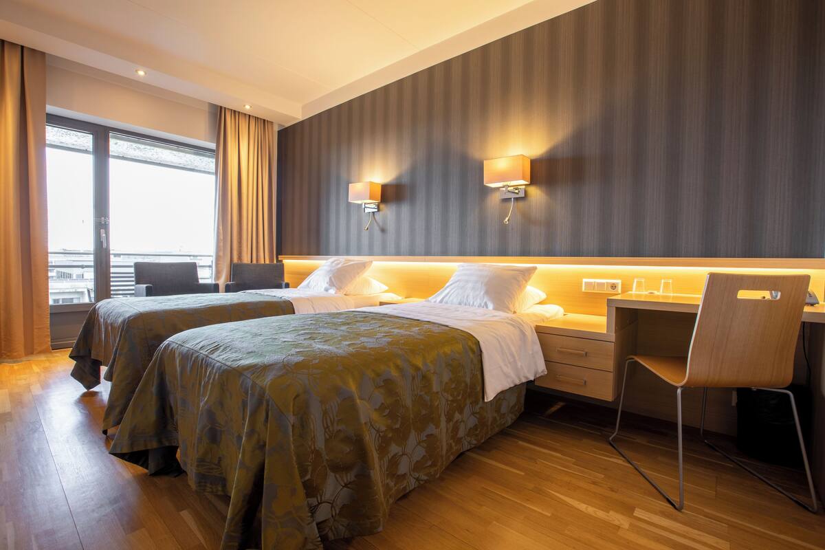 A hotel room with two beds, a desk, a chair, and a view of the city.