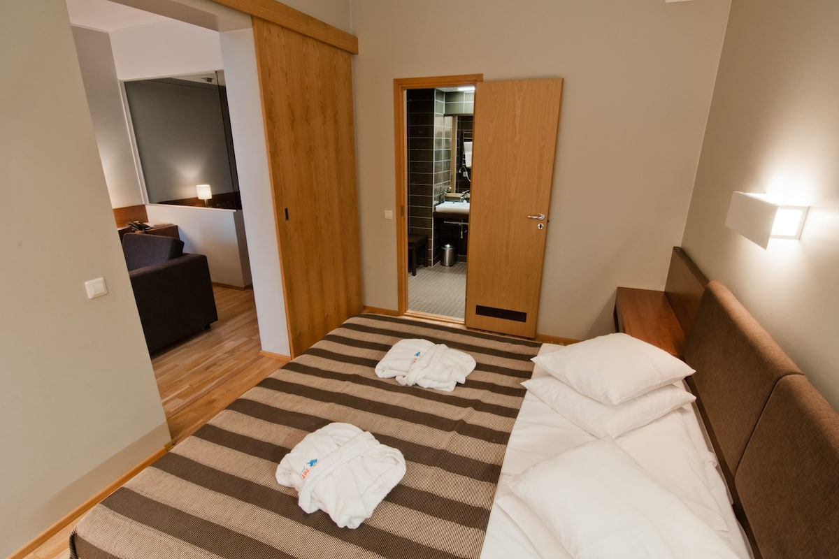 A hotel room with a bed, a wooden wardrobe, a mirror, and a bathroom visible through an open door.
