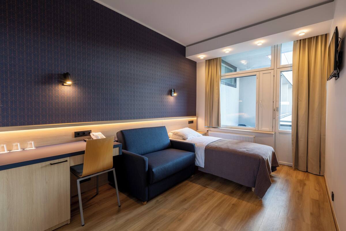 A hotel room with a desk, a chair, a bed, a window with curtains, and a wall-mounted TV.