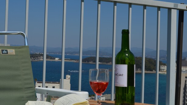Un balcon avec vue sur la mer, une bouteille de vin verte, un verre de vin rouge et un livre ouvert.