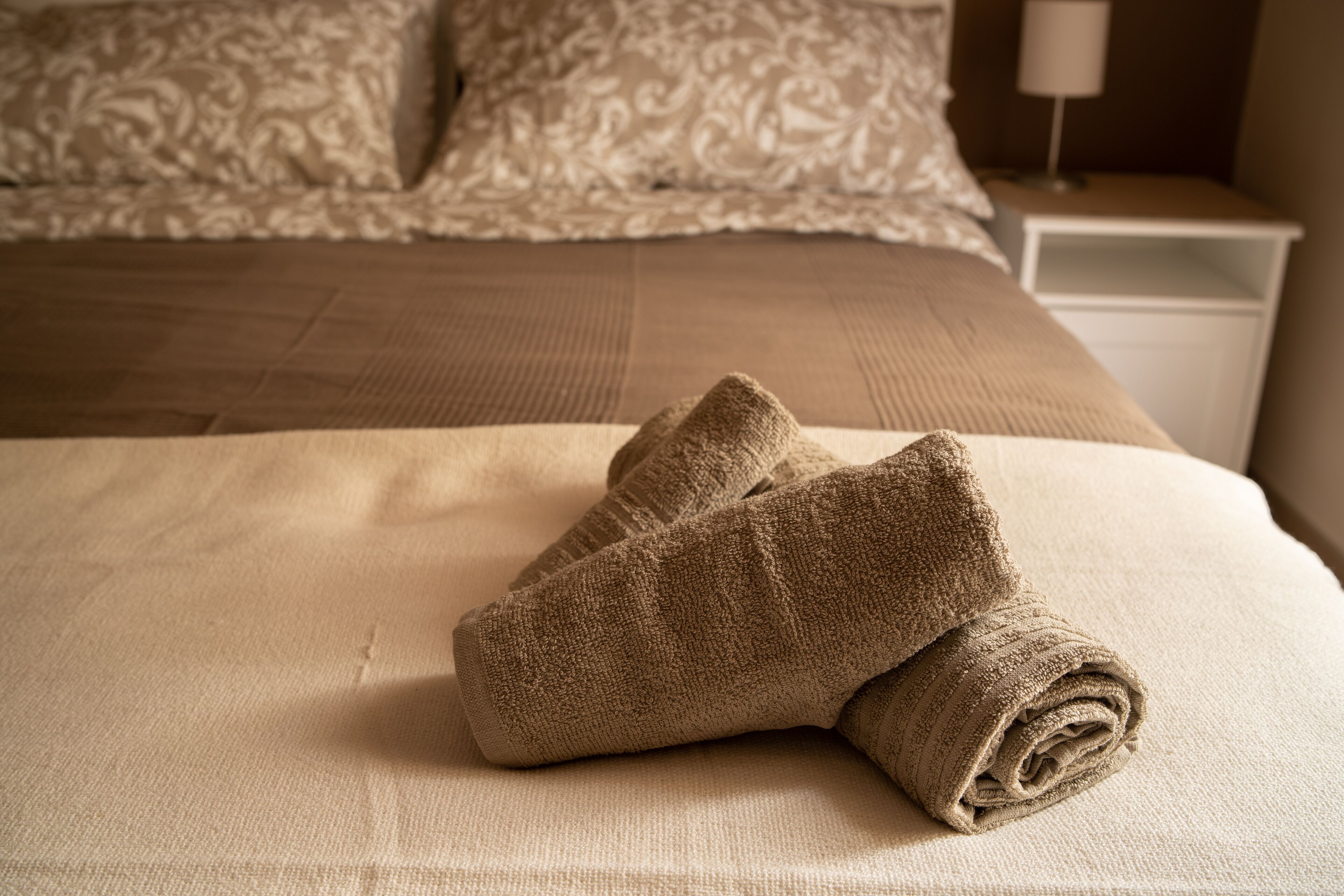 Un letto ben rifatto con copripiumino color beige e cuscini, un comodino bianco con una lampada, e un asciugamano arrotolato sul letto.