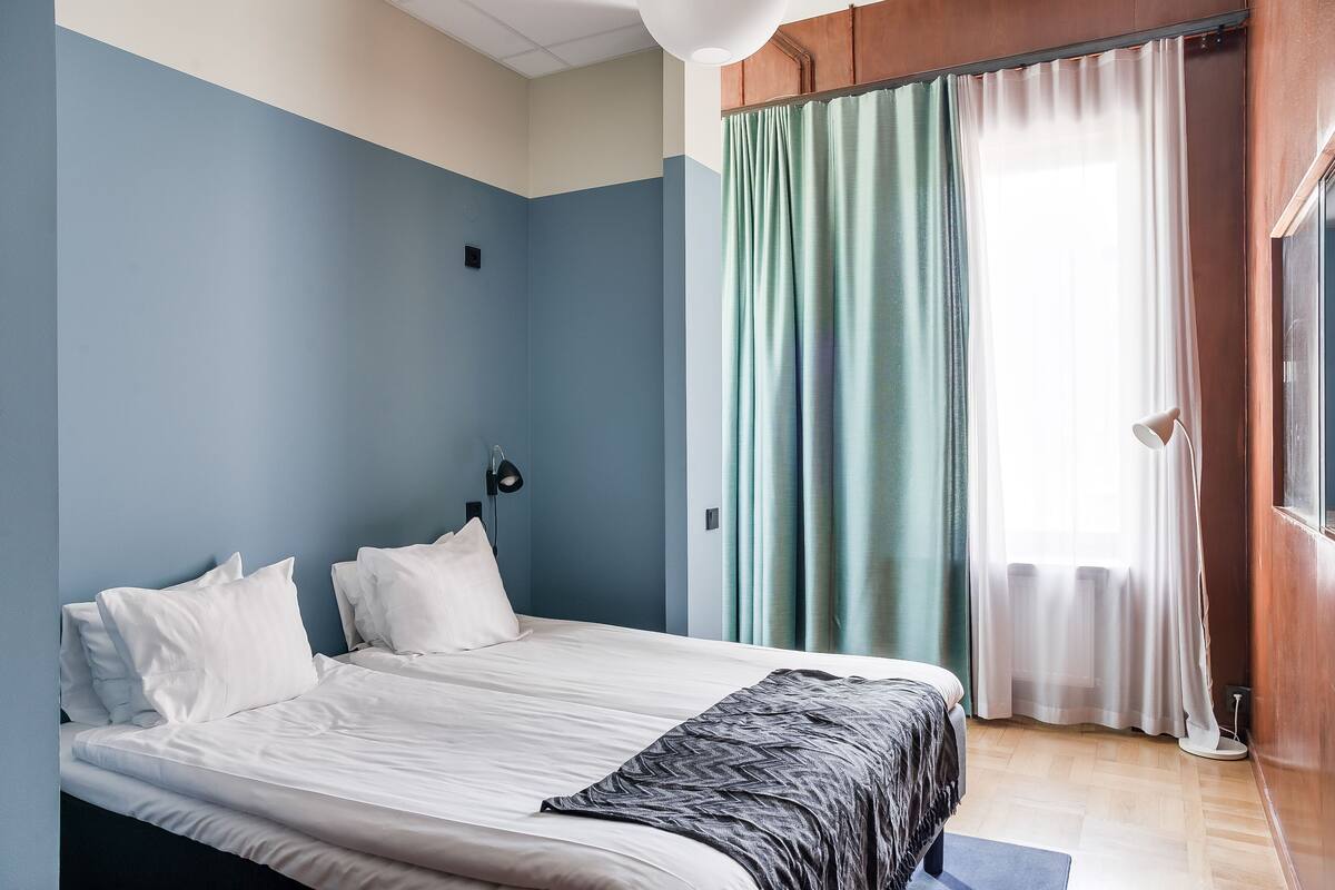Un lit bien fait, avec du linge de lit blanc et un plaid gris, une lampe de chevet et une vue sur une fenêtre avec des rideaux.