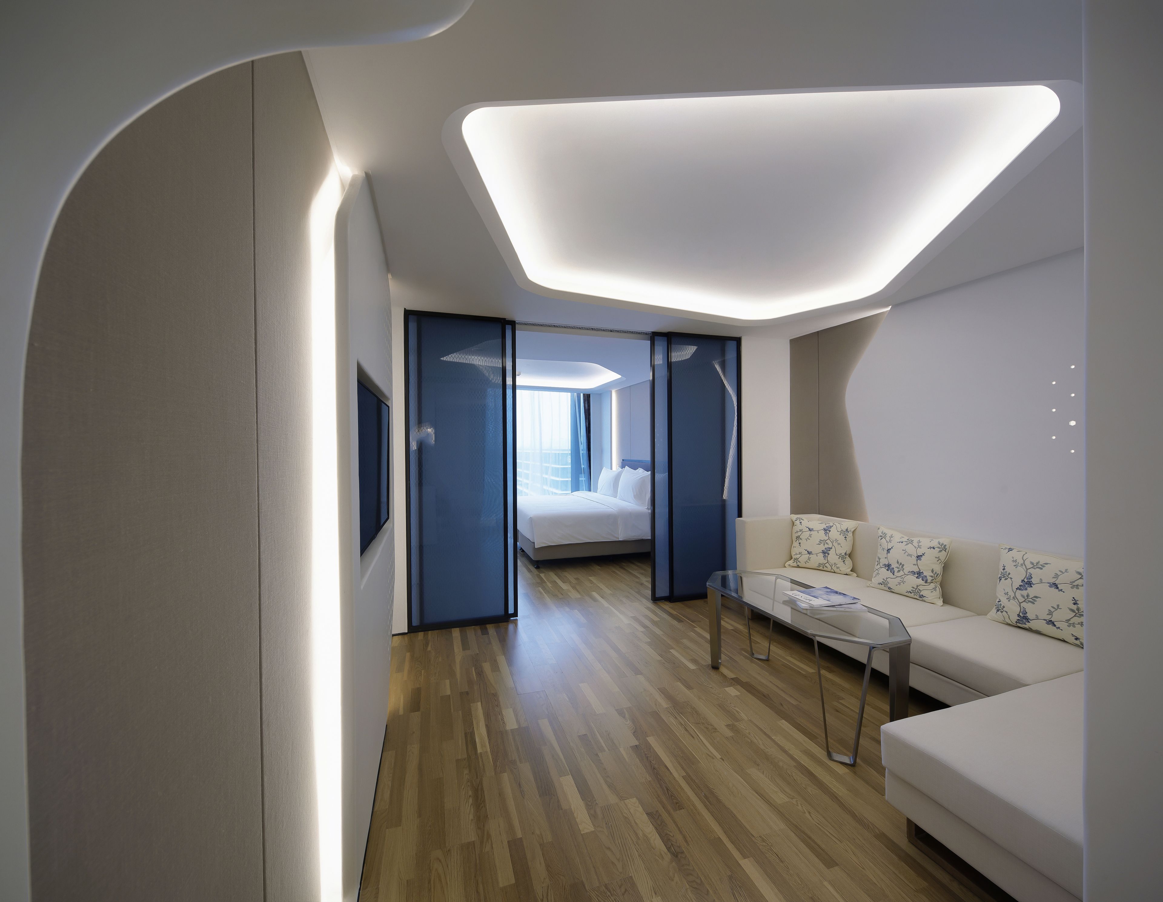 Una habitación de hotel moderna con piso de madera, un sofá blanco, una mesa de centro de vidrio y una cama con ropa blanca de cama.