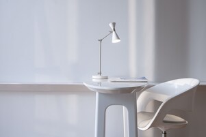 Una habitación moderna y minimalista con un escritorio y una silla blancos, una lámpara de pie blanca y un ventanal que deja entrar luz natural.