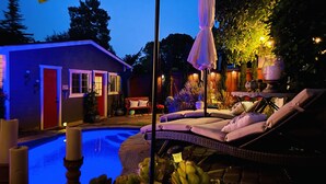 Una piscina con sillones, una puerta roja y un jardín con plantas y una mesa.
