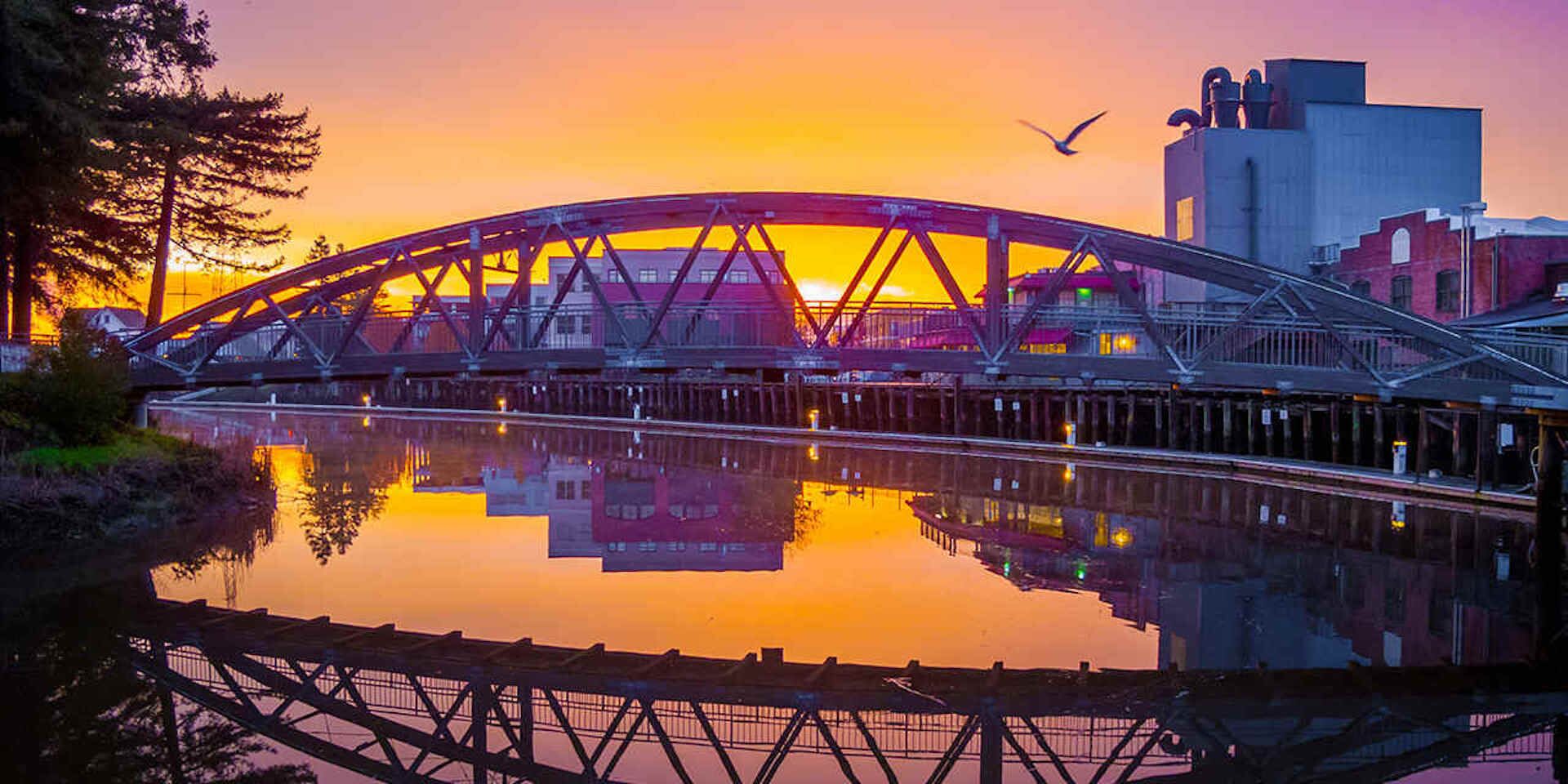Un pont d’acier enjambant une étendue d’eau calme, avec un coucher de soleil en arrière-plan.
