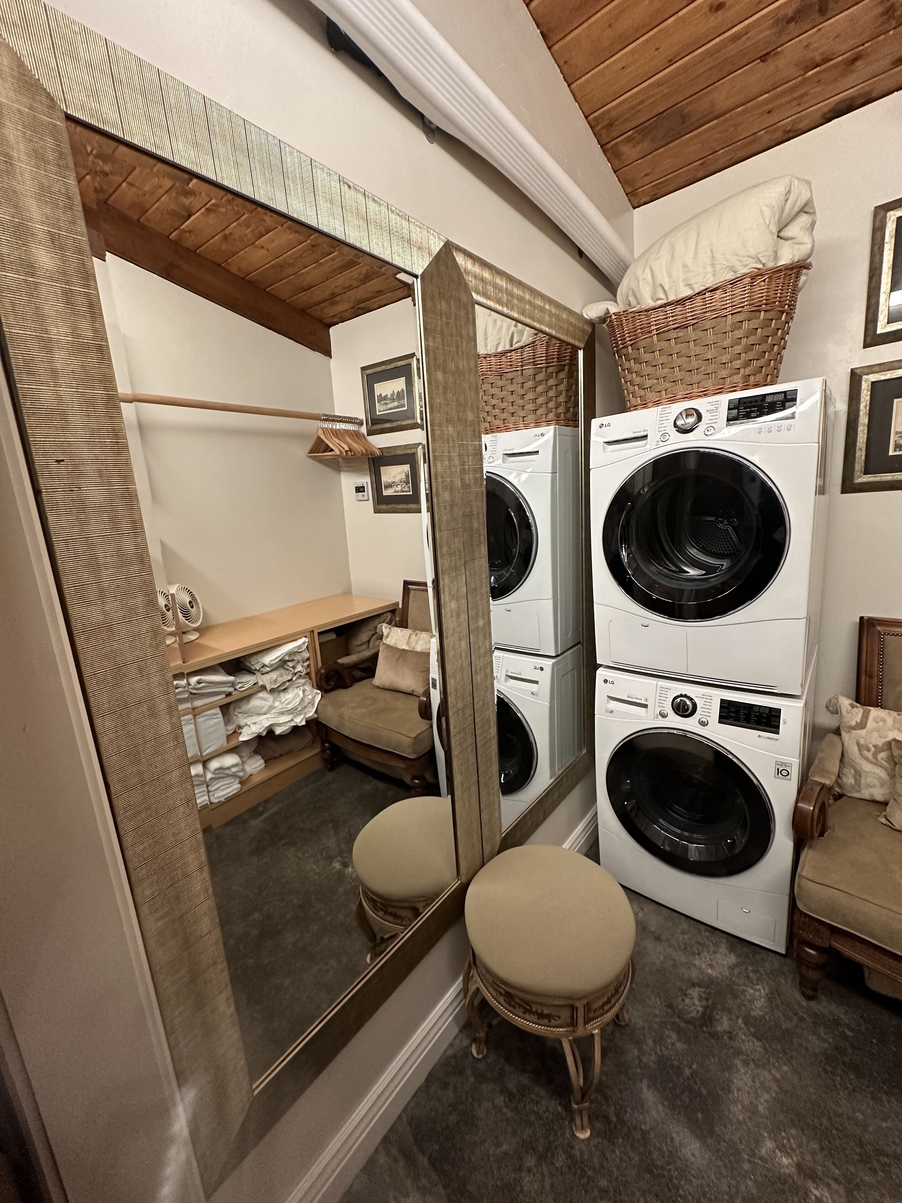 Une petite buanderie comprenant deux machines à laver à chargement frontal superposées, un miroir, un panier en osier posé sur les machines, une chaise en bois et un petit tabouret.