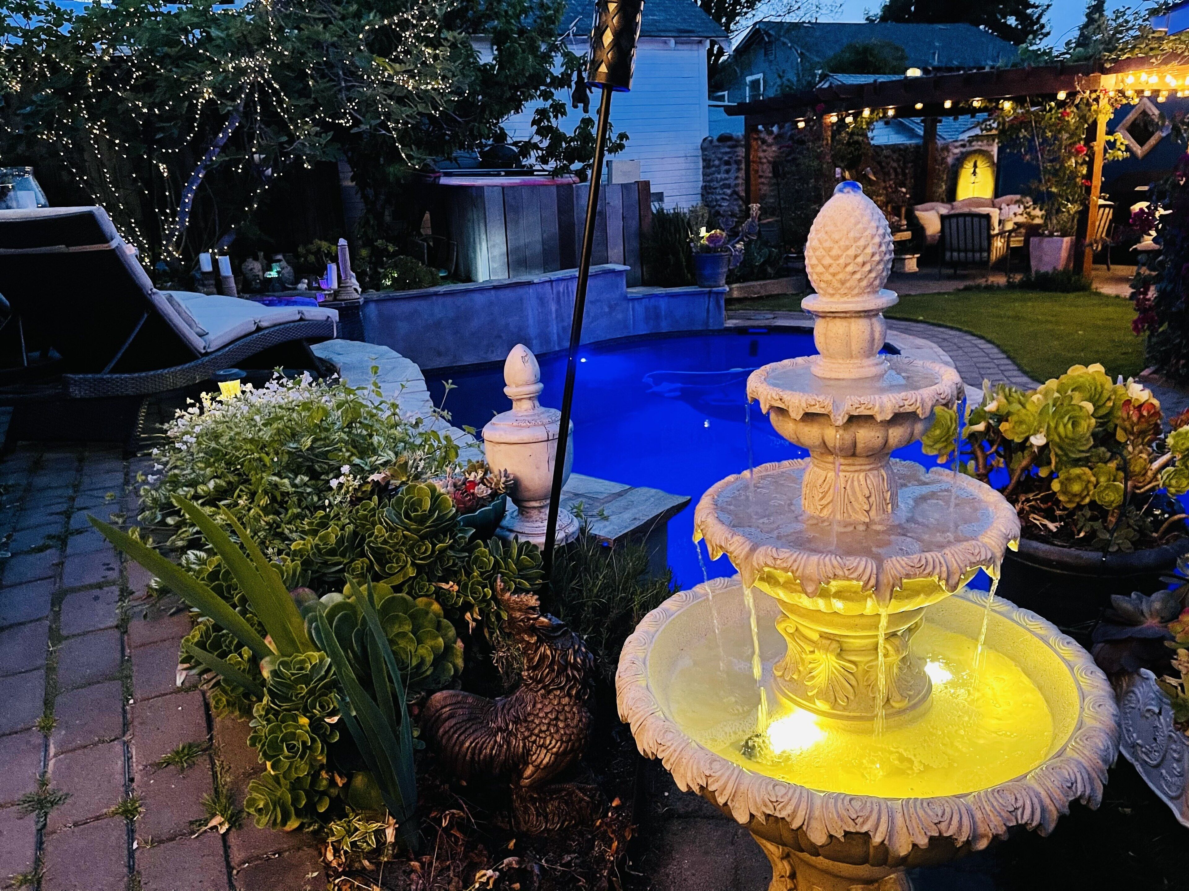 A multi-tiered stone fountain with water flowing, surrounded by lush greenery and potted plants.