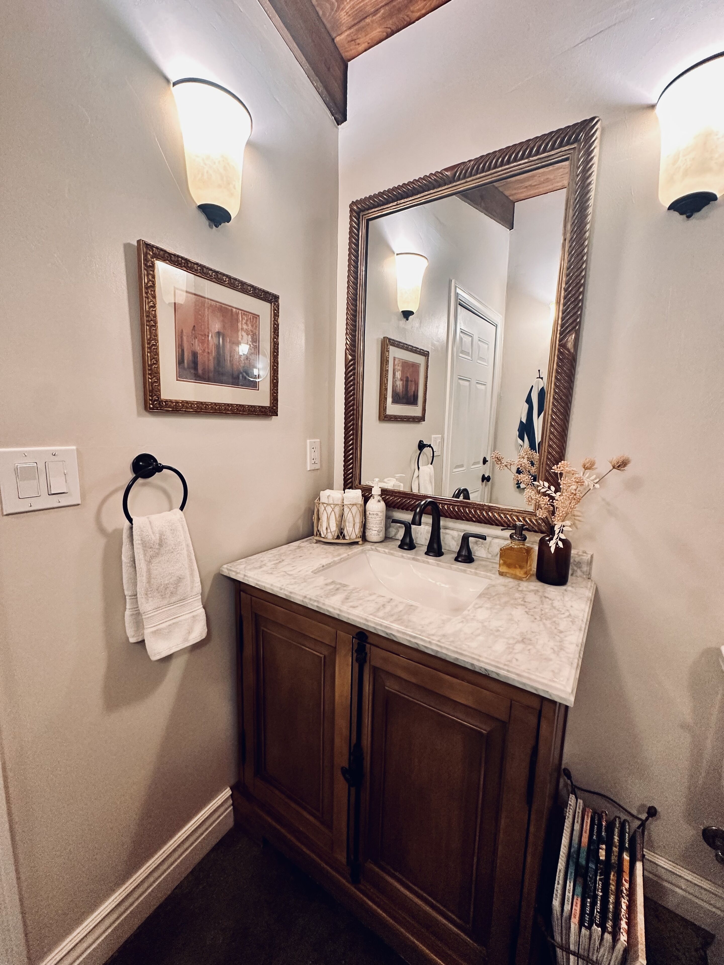 A bathroom with a marble sink, wooden cabinets, a large mirror, wall sconces, a towel rack, and framed pictures.