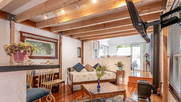A cozy room with a bed, a wooden table, a chair, a rug, and a window with plants.