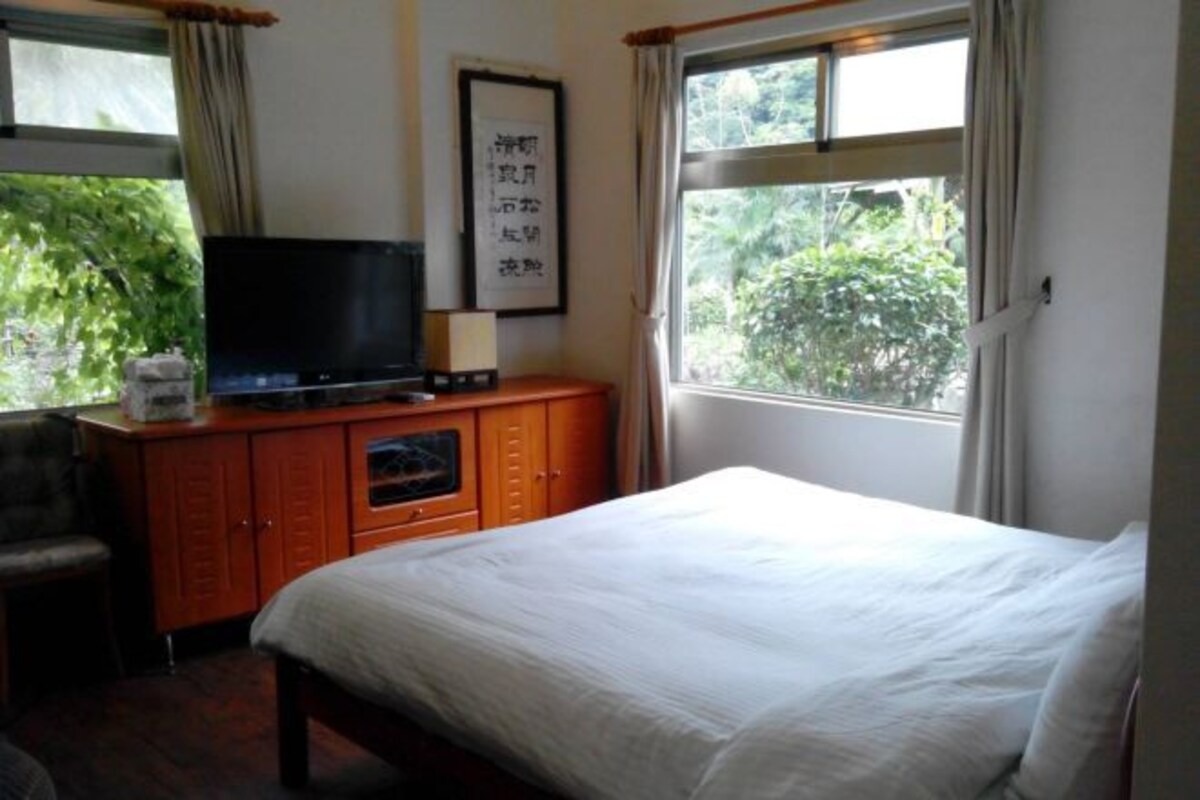 A hotel room with a bed, a television on a wooden cabinet, a desk, and a window with a view of greenery.