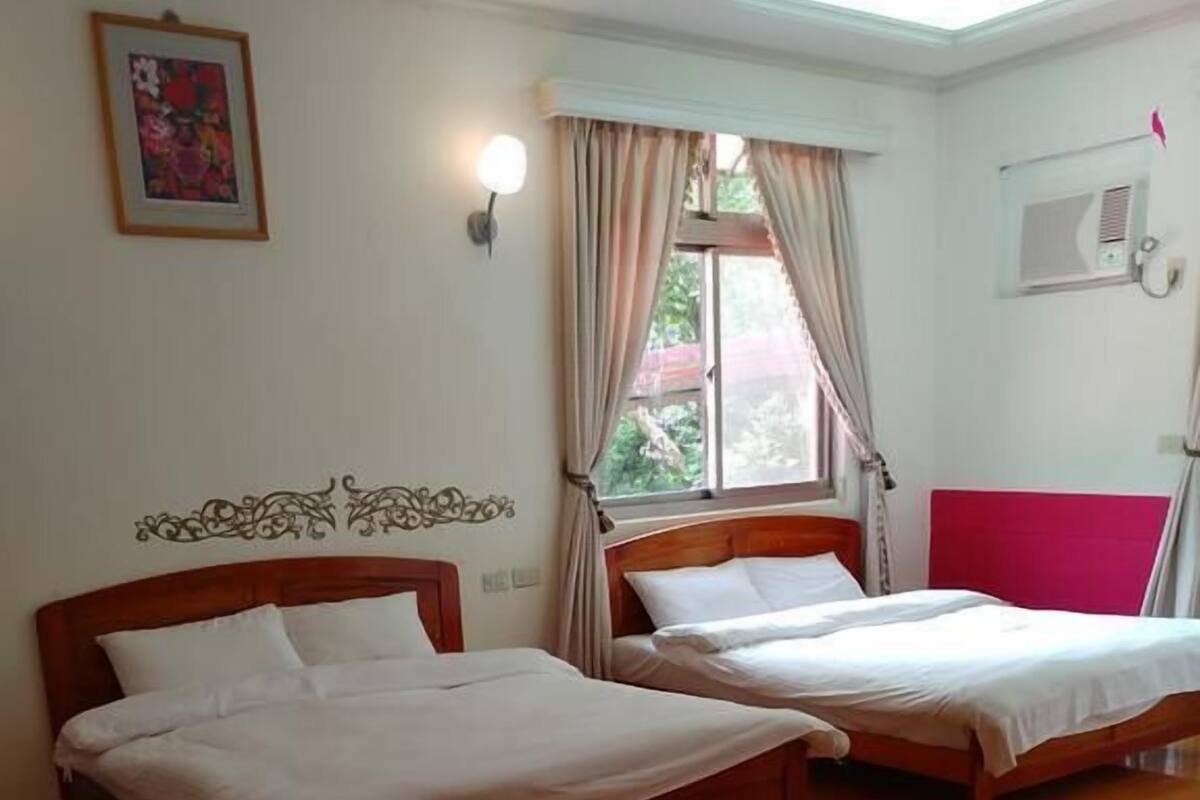 A hotel room with two wooden beds, a window with curtains, a wall-mounted air conditioning unit, and a decorative wall piece.