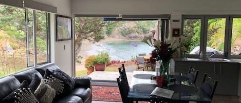 A modern living room with a black leather sofa, a glass dining table, and a view of a beach through large windows.