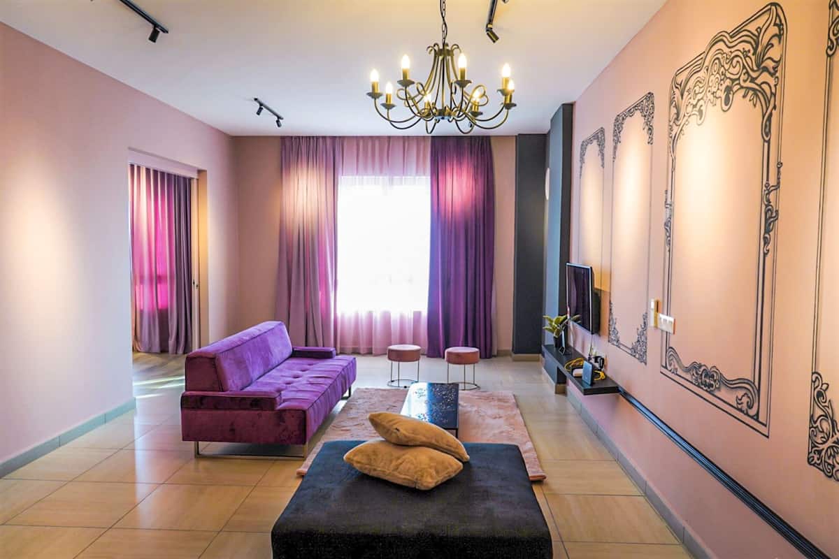 Uma sala de estar moderna com um sofá roxo, um banquinho preto com almofadas, um candelabro e um espelho grande com detalhes ornamentados.