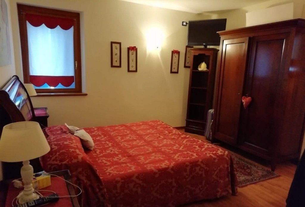 A bedroom with a bed, a nightstand, a lamp, a TV, a wardrobe, and a window with curtains.
