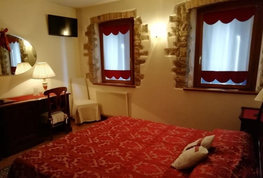 A bedroom with a red patterned bedspread, a chair, a desk with a lamp, and two windows with red curtains.