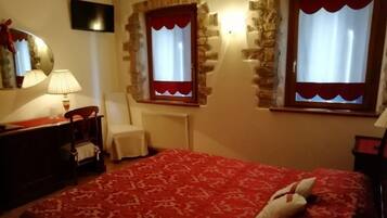 A bedroom with a red patterned bedspread, a chair, a desk with a lamp, and two windows with red curtains.
