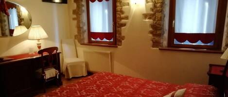 Une chambre avec une couvre-lit à motifs rouges, une chaise, un bureau avec une lampe et deux fenêtres ornées de rideaux rouges.