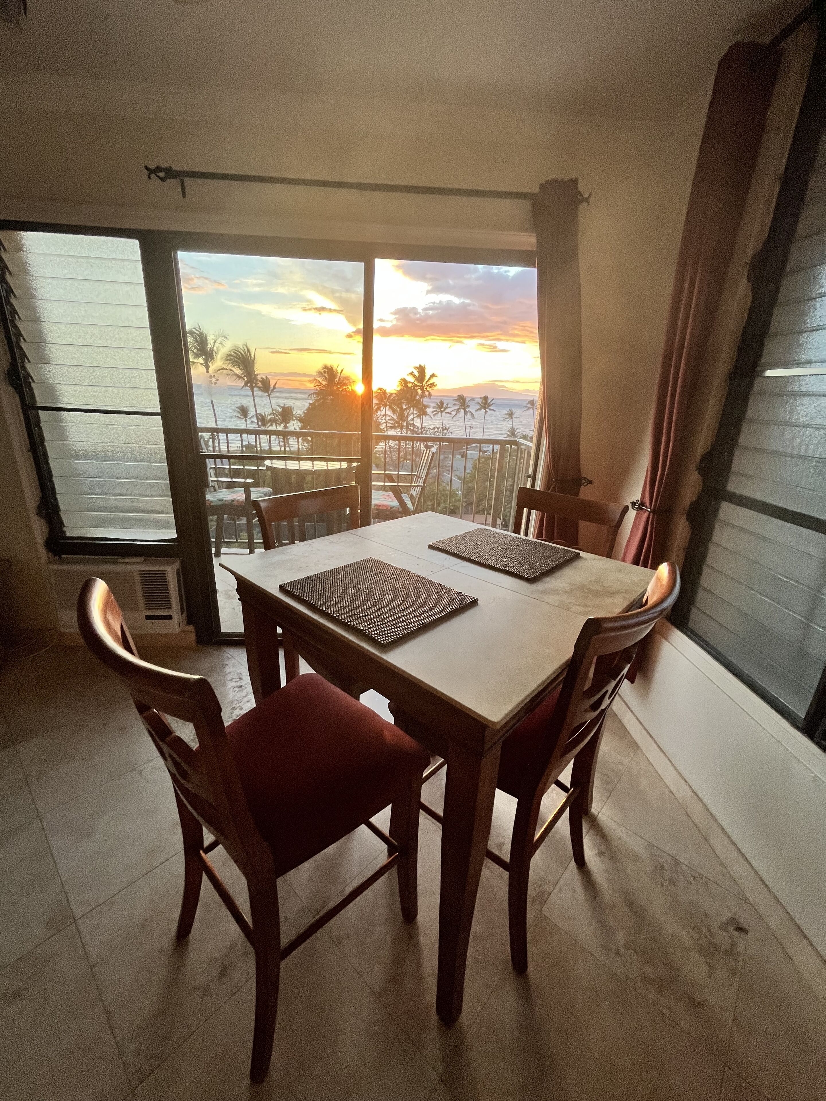A dining area with a table and chairs, a view of the sunset over the ocean, and a balcony with a railing.