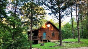 A rustic log cabin with a porch and a small deck, surrounded by dense forest.