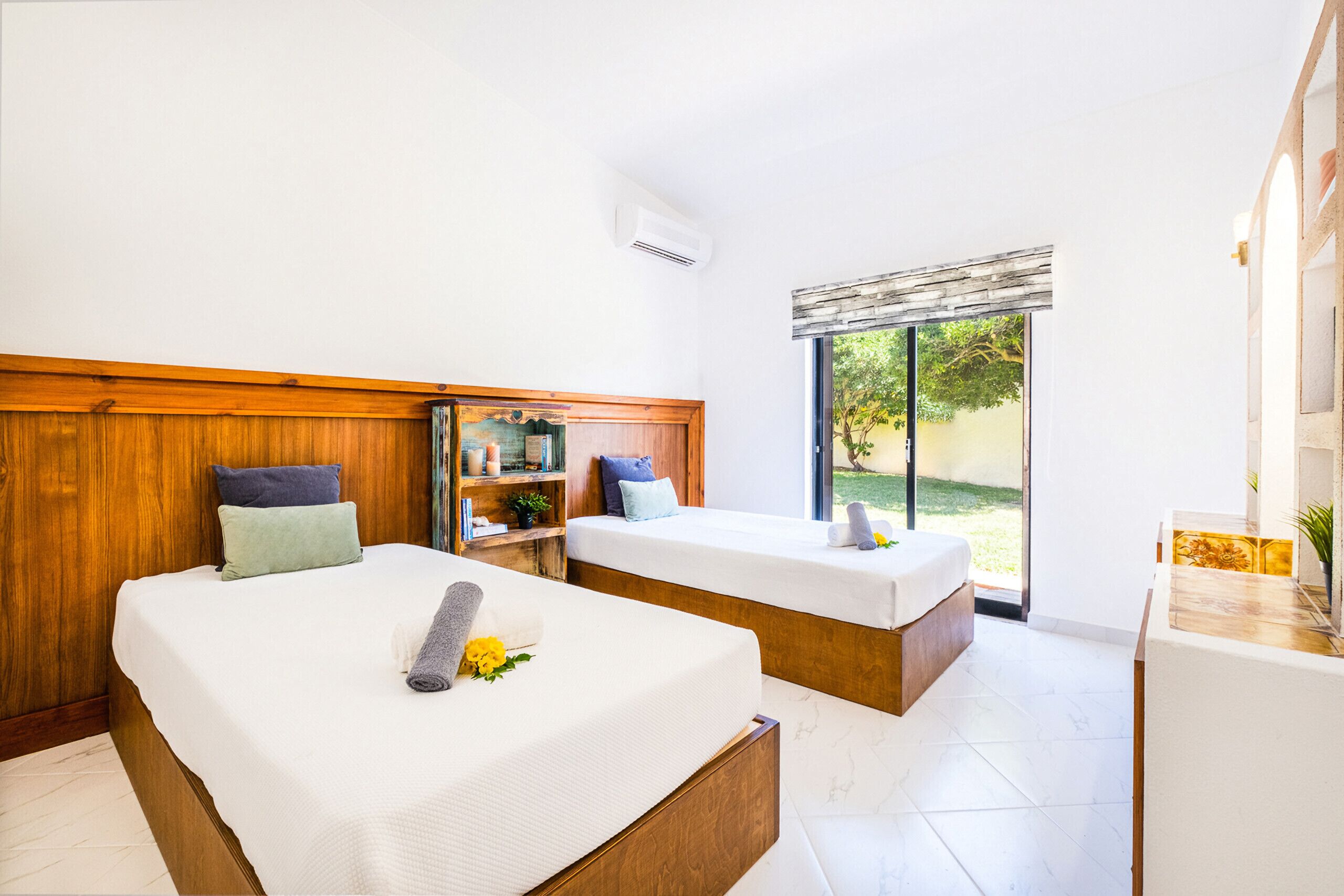 Une chambre d’hôtel avec deux lits, une tête de lit en bois, un petit bureau, une fenêtre donnant sur un espace verdoyant et une salle de bain avec un miroir et un lavabo.