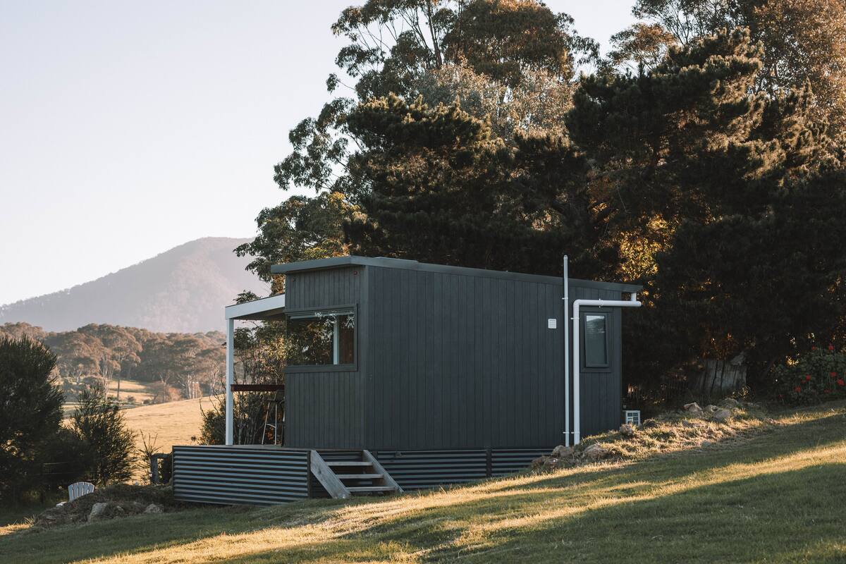 A modern, single-story building with a flat roof, situated on a grassy hill with a backdrop of trees and a mountain.