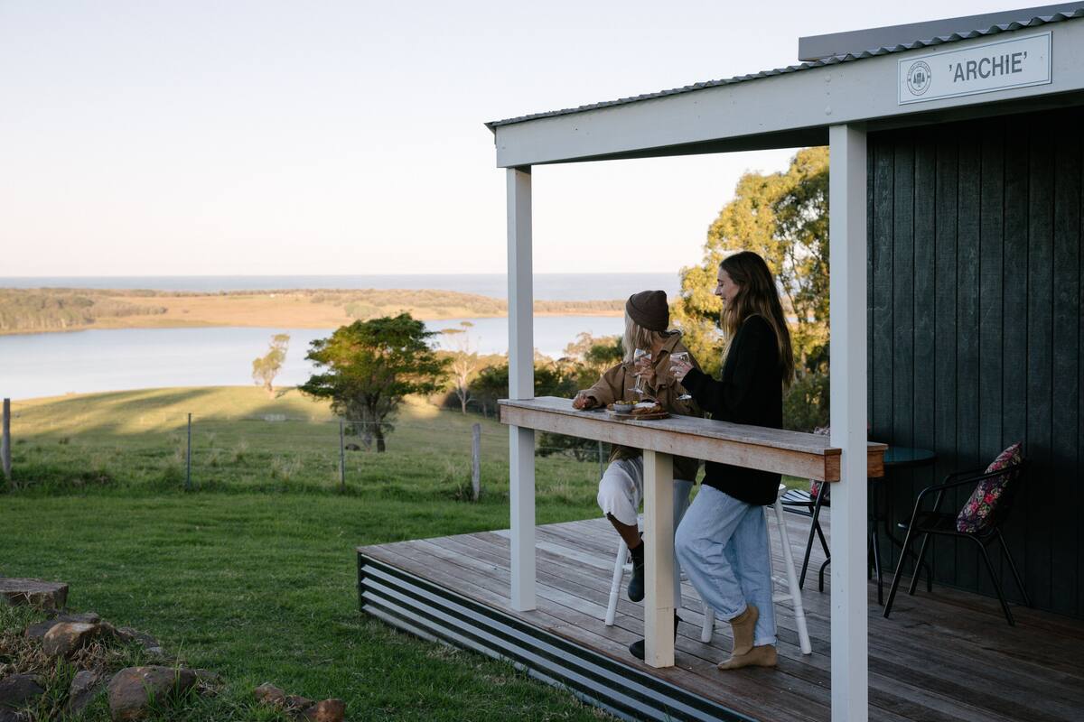 A wooden deck with a white railing, a sign reading 'ARCHIE', and a view of a lake and trees in the background.