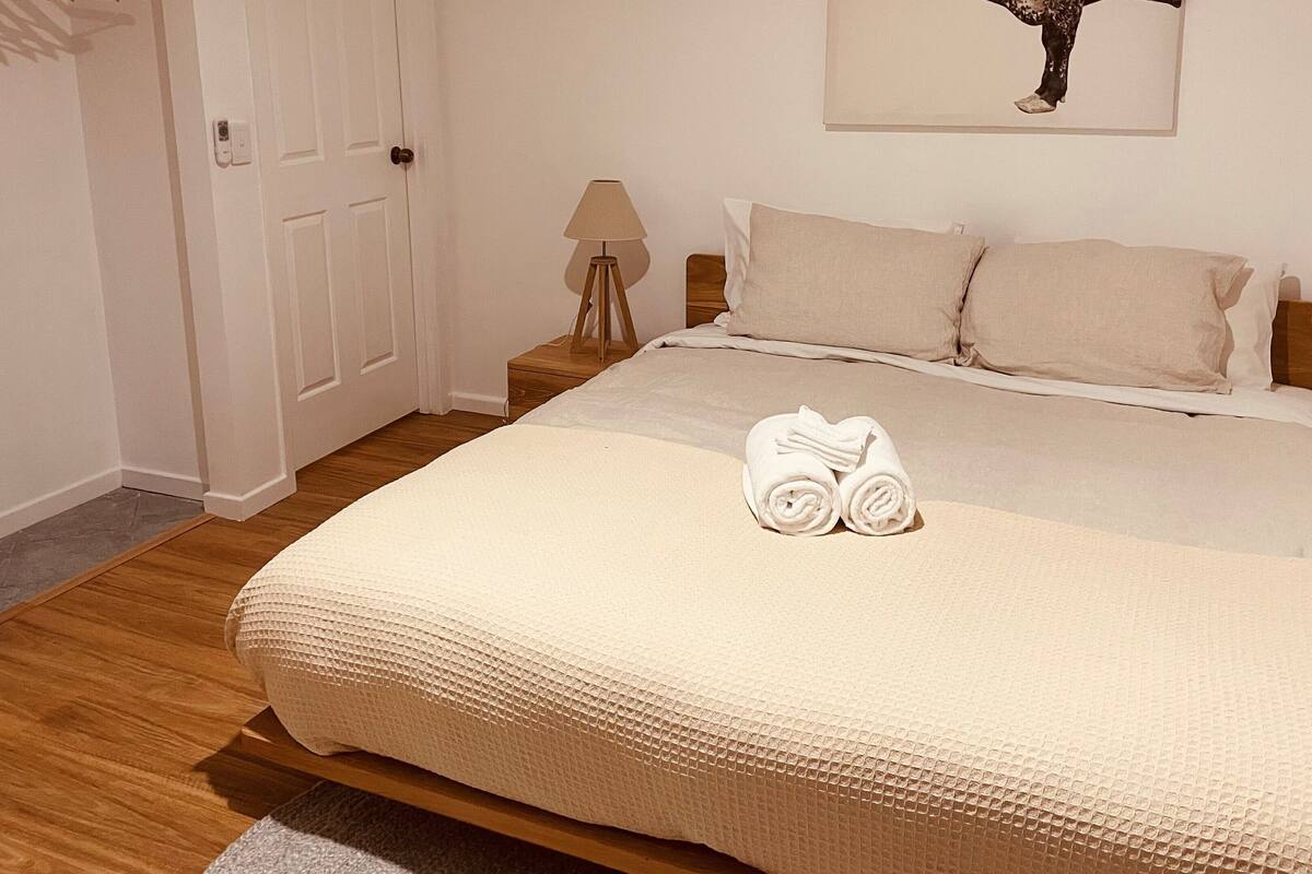 A neatly made bed with a rolled towel, a bedside lamp, and a wooden floor.