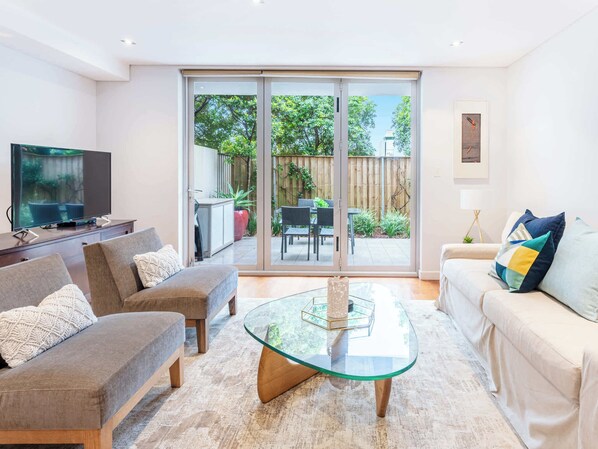 A living room with a TV, sofas, a glass coffee table, and a sliding glass door leading to an outdoor patio.