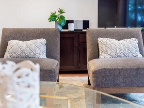 A modern living room with two grey armchairs, a dark wooden cabinet, a potted plant, and a glass coffee table.