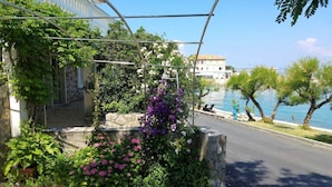 Une rue bordée d’un mur de pierre, une pergola ornée de fleurs et une vue sur une étendue d’eau, avec des bâtiments en arrière-plan.