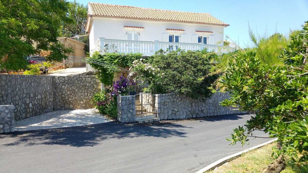 A two-story building with a stone wall and a paved driveway leading to a gated entrance.