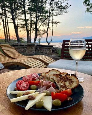 A plate of food with a glass of white wine, set on a table outdoors with a view of trees and a sunset. - Apartment Sanjug (Silo - island Krk)
