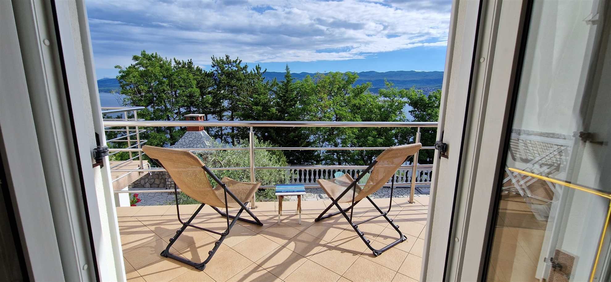 A balcony with two wicker chairs, a small table, and a view of a lake and mountains.