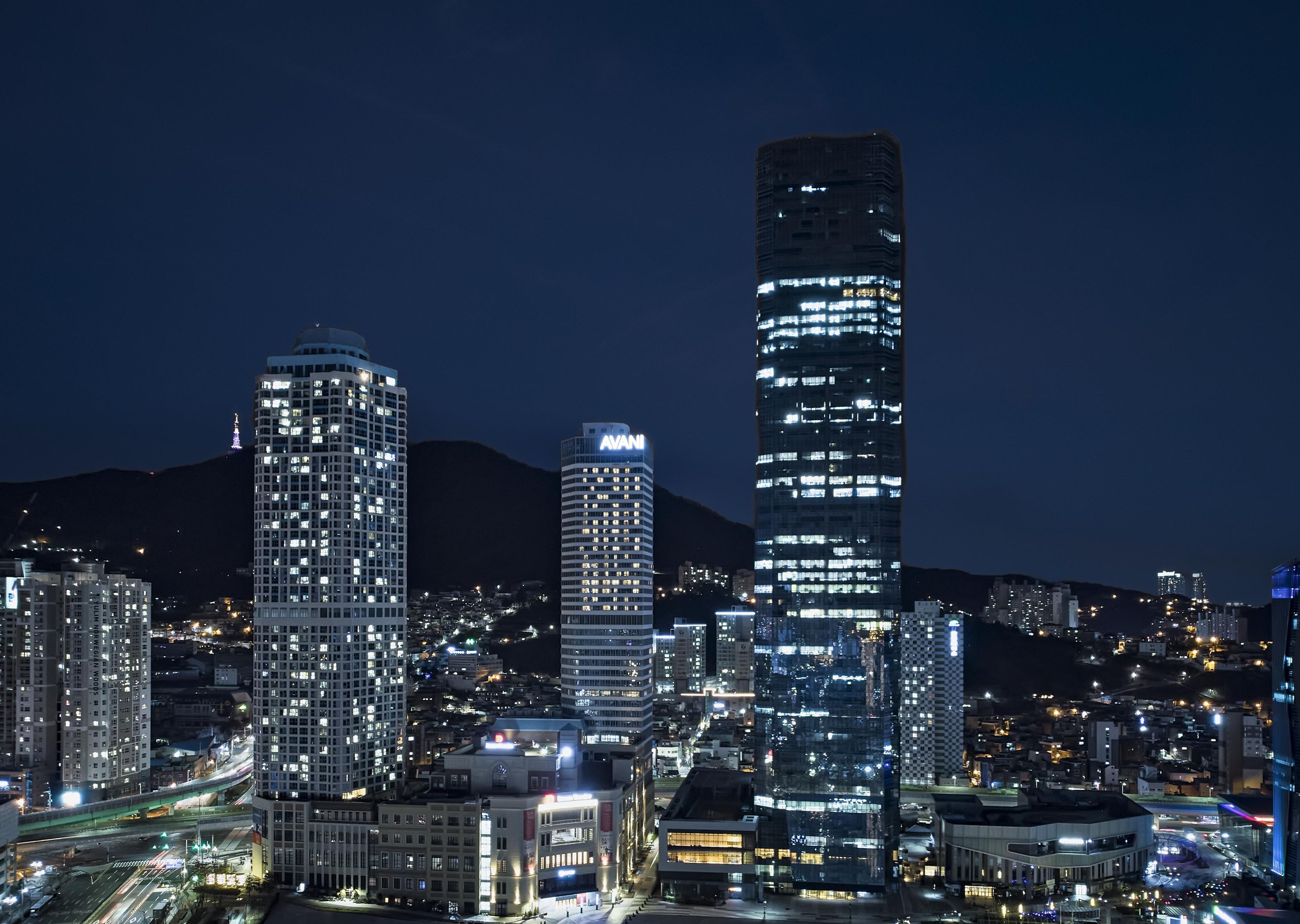 A nighttime cityscape with illuminated skyscrapers, including a prominent building with 'AVANI' signage.