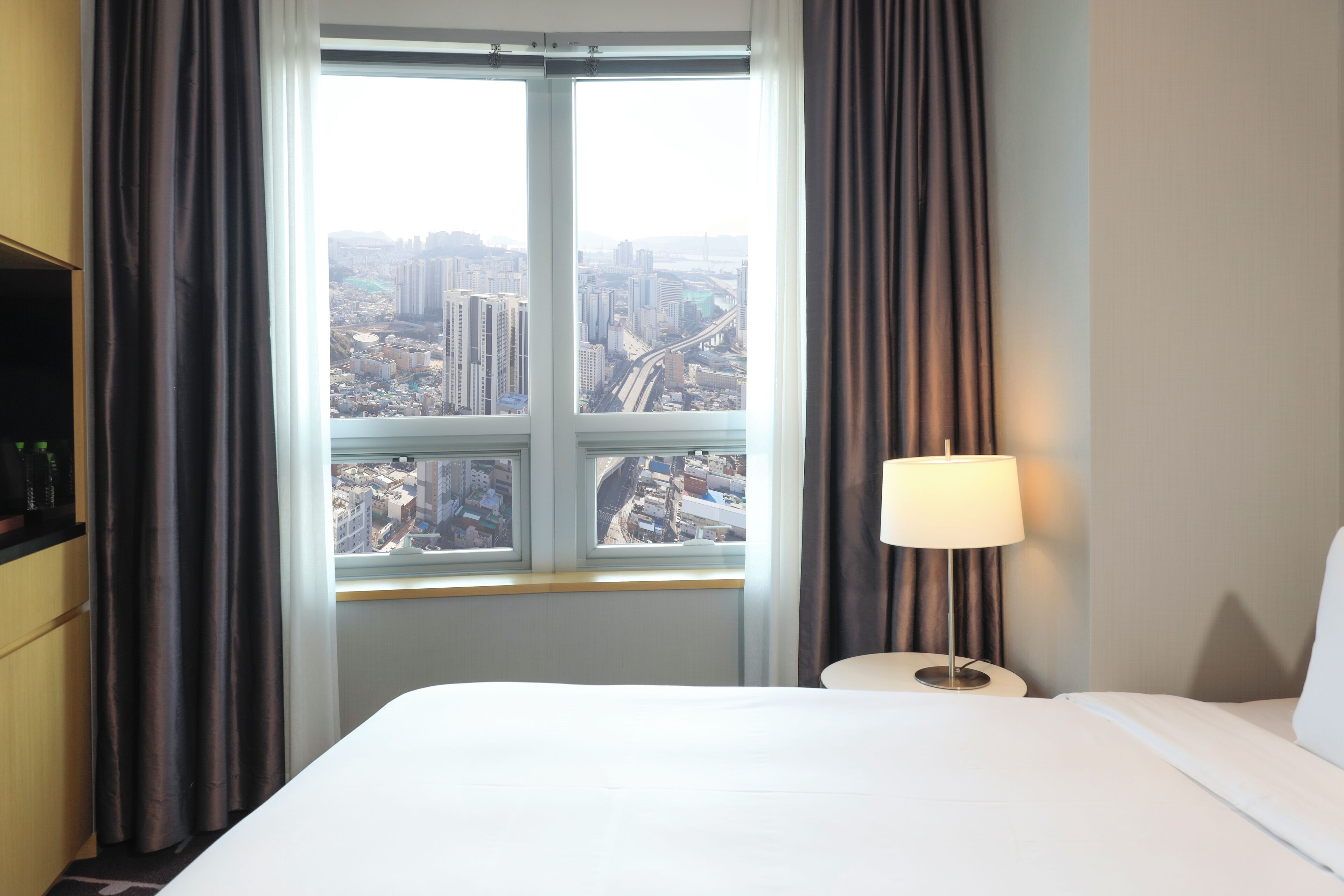 A hotel room with a large window offering a city view, a bed, a bedside table with a lamp, and a TV on the left side.