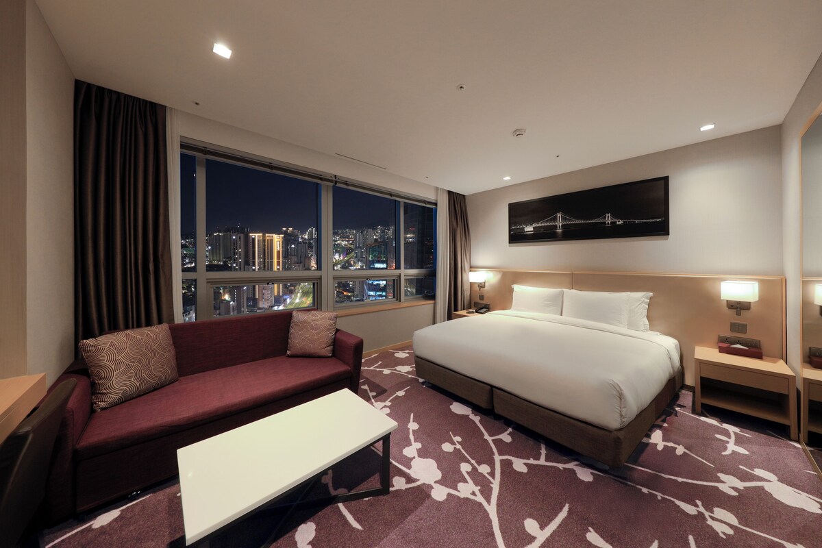 A hotel room with a large bed, a sofa, a coffee table, and a view of the city at night.