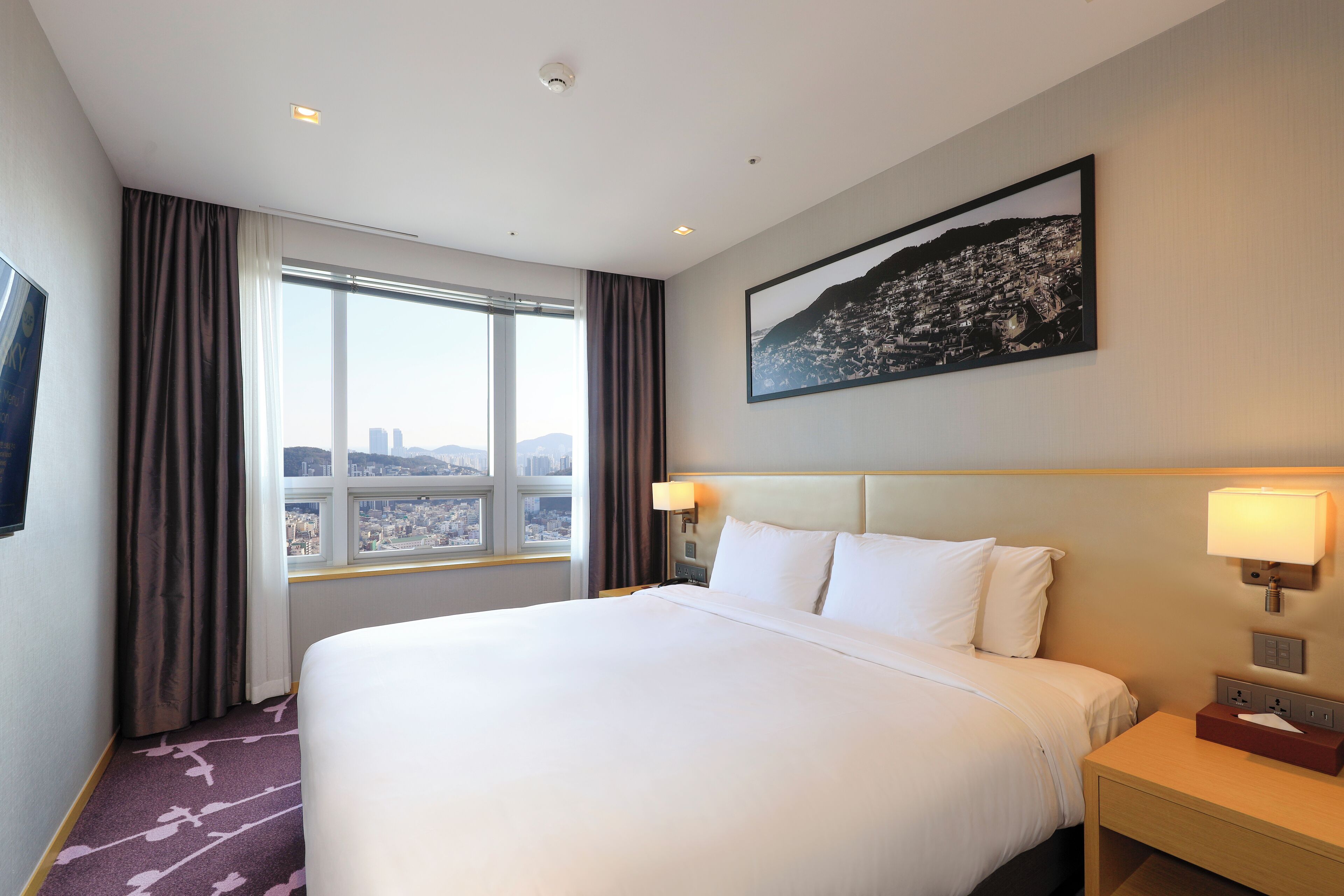 A hotel room with a large bed, two bedside lamps, a nightstand, a window with curtains, and a cityscape view.