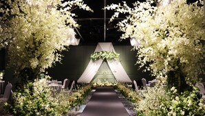 A long, narrow indoor space with a central aisle, flanked by white floral arrangements and a draped canopy. Tables and chairs are arranged on either side of the aisle. - Avani Central Busan (Busan)