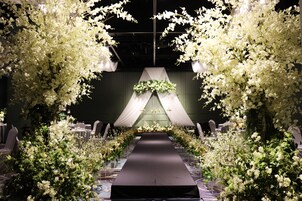Un espacio interior largo y estrecho con un pasillo central, flanqueado por arreglos florales blancos y un dosel colgante. Mesas y sillas están dispuestas a ambos lados del pasillo.