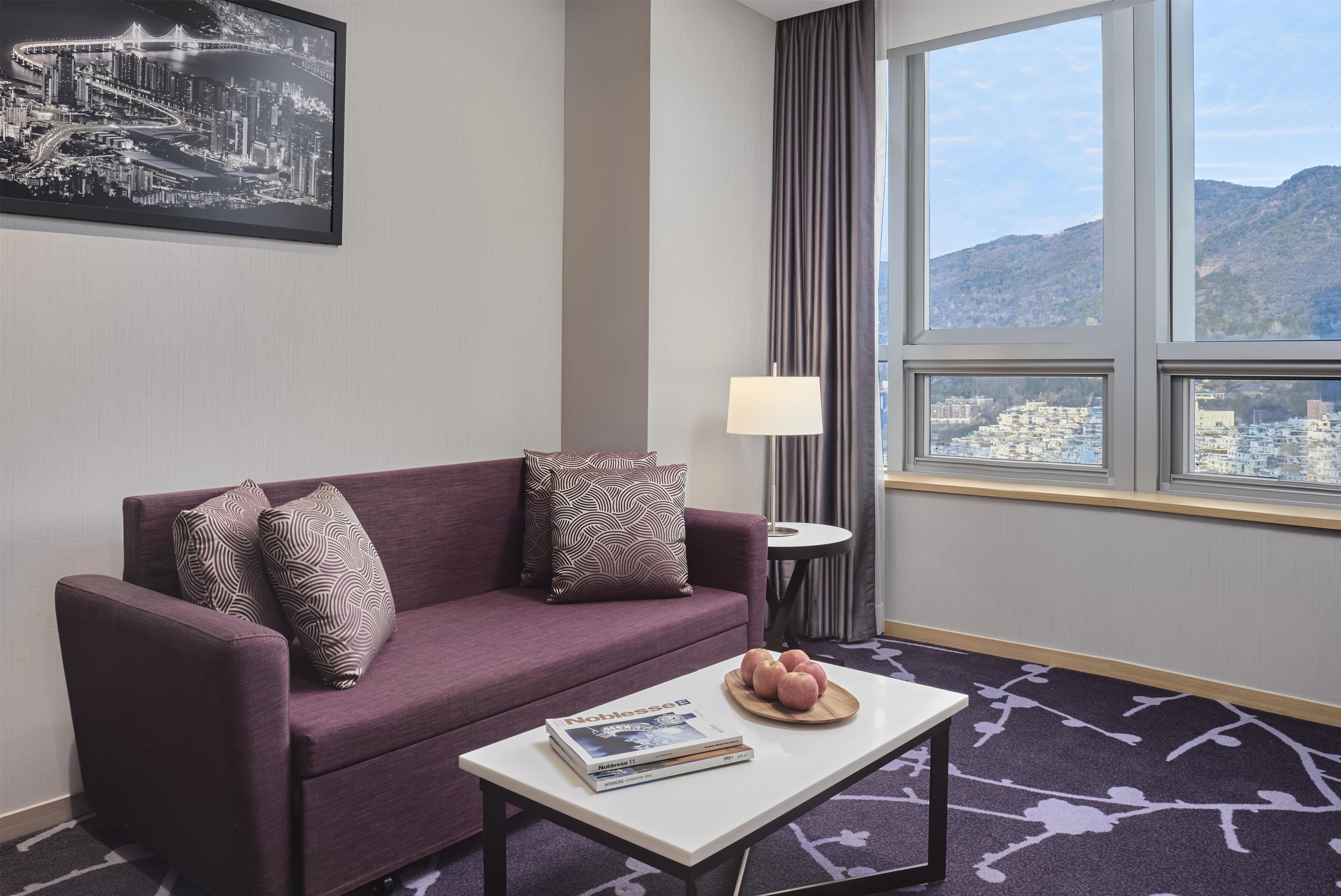 Habitación de hotel con sofá morado, almohadas estampadas, una mesita con una revista, una lámpara y vistas de la ciudad a través de un ventanal grande.