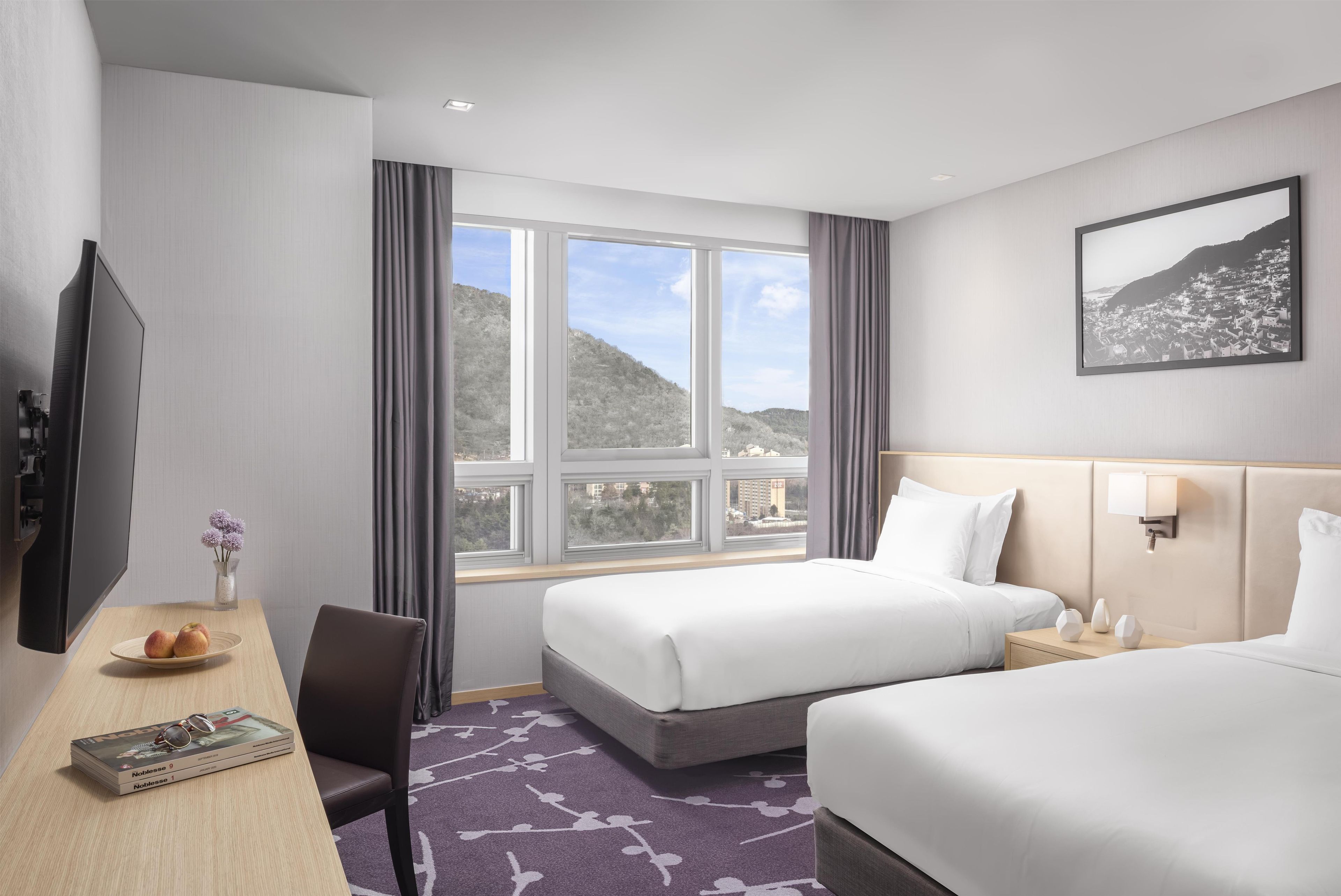 Habitación de hotel con dos camas, un escritorio con televisor, una ventana con vista a un paisaje urbano y un cuadro en la pared.