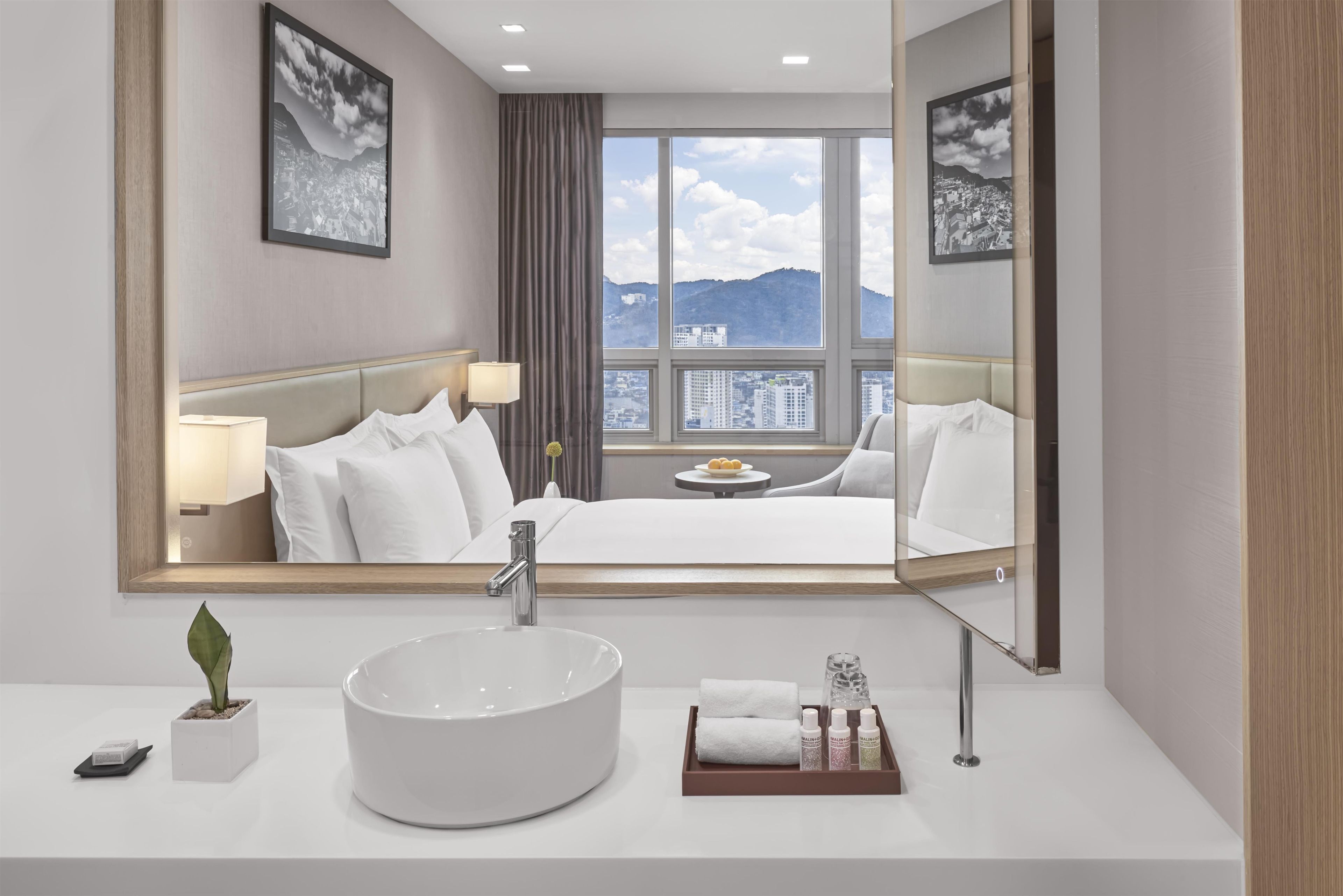 Habitación de hotel con una cama grande, un lavabo redondo blanco, un espejo y vistas a un paisaje urbano a través de un ventanal amplio.