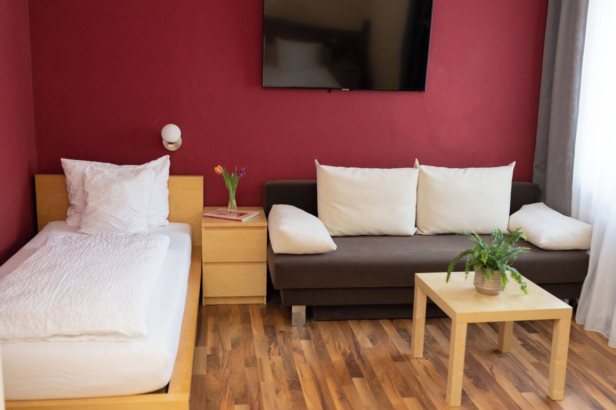 Una habitación de hotel con cama, sofá, una mesita con una planta, mesita de noche y televisor en la pared.