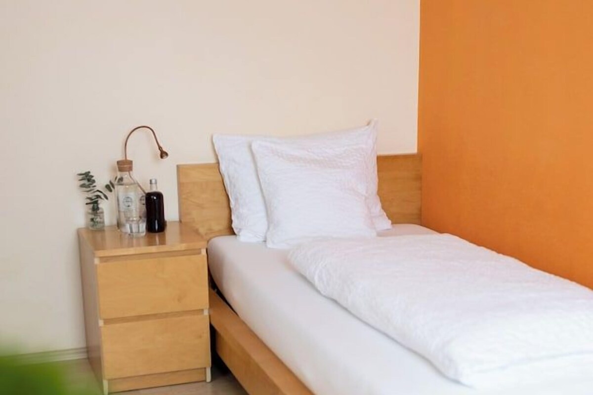 Una cama individual con estructura de madera, una mesita de noche con cajones y una botella de agua sobre la mesita.