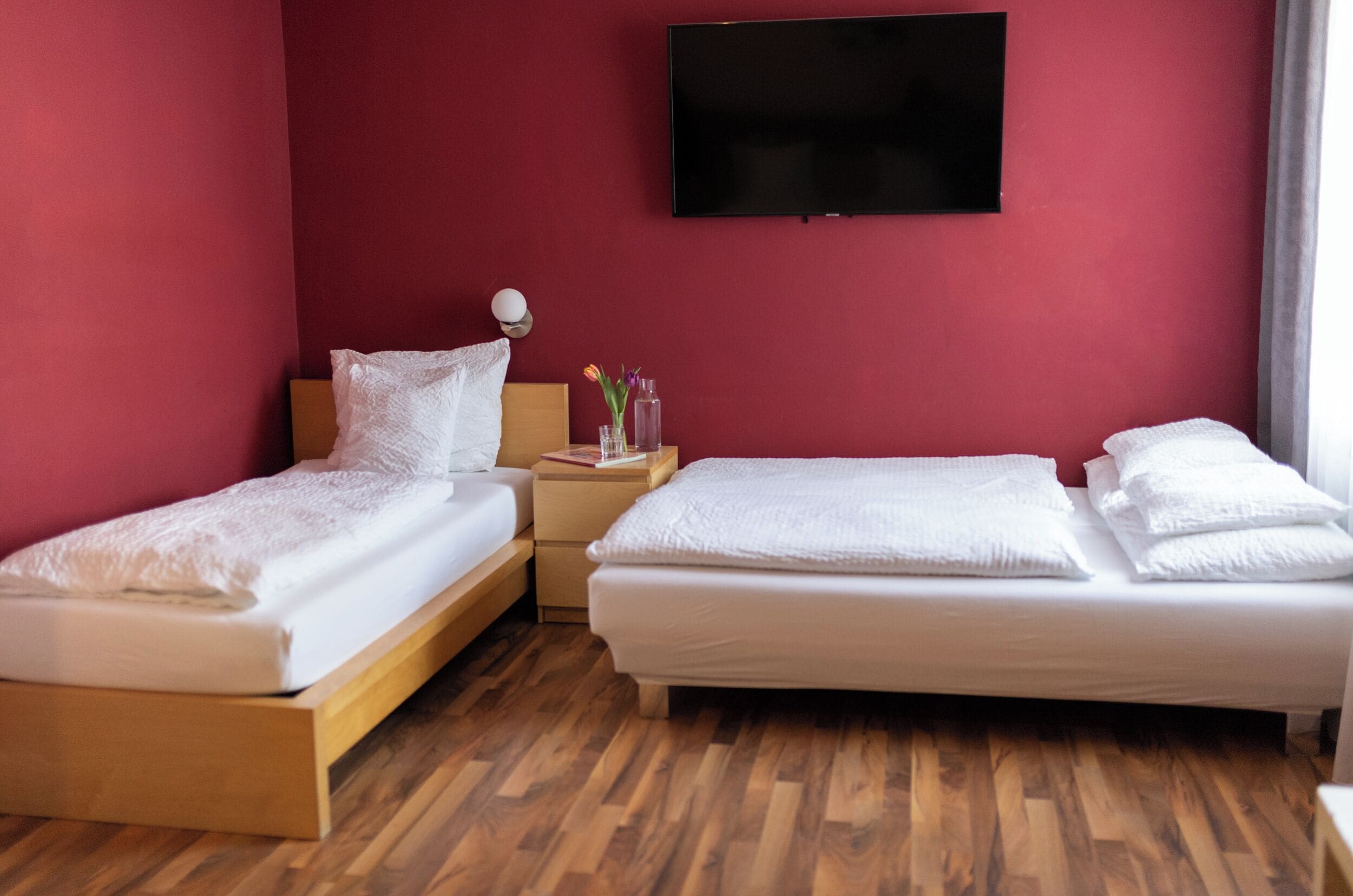 Une chambre d’hôtel avec deux lits simples, un plancher en bois, un mur rouge, un téléviseur à écran plat et une table de chevet avec une lampe et un vase de fleurs.