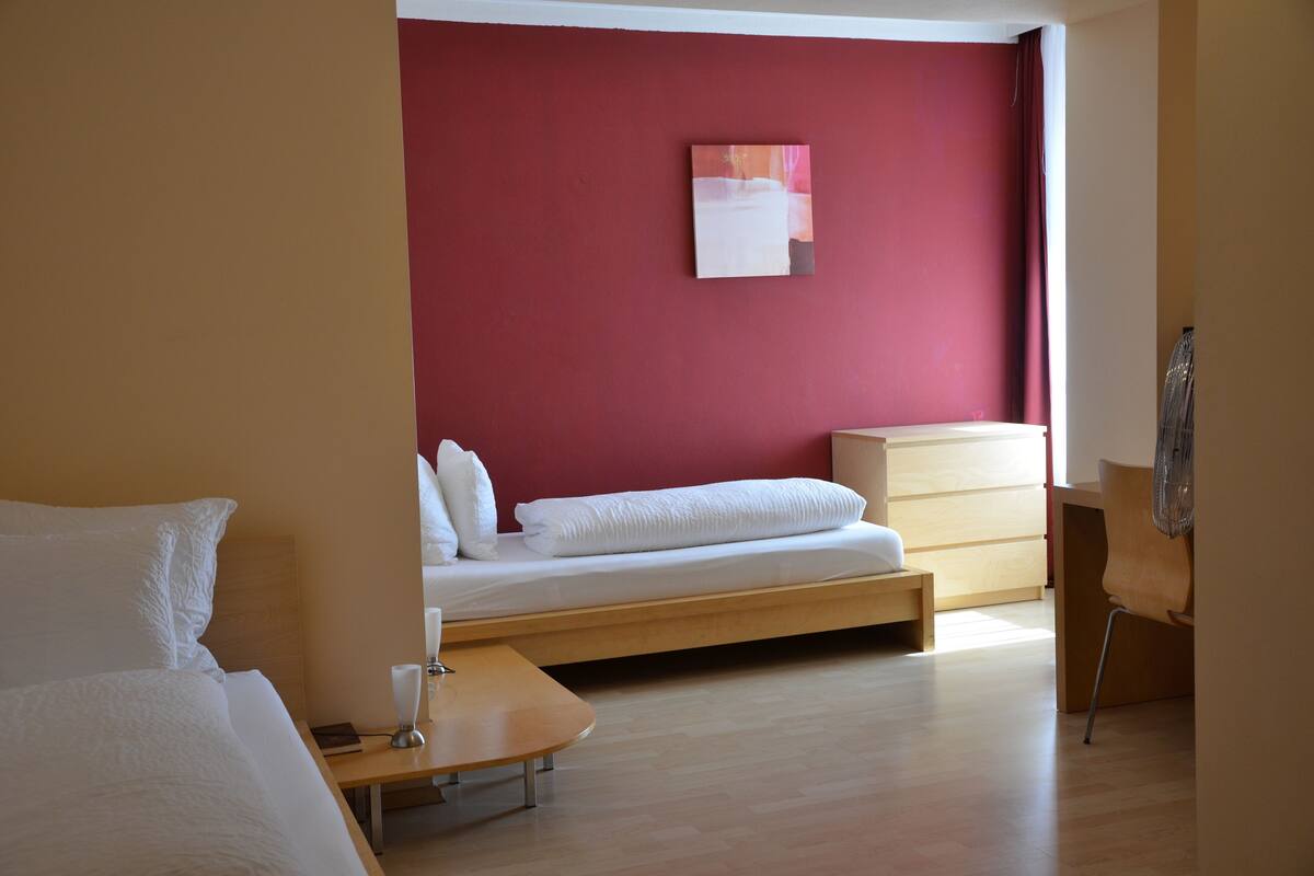 Una habitación de hotel con una cama, una mesita de noche de madera, una silla y una pared roja con un cuadro.
