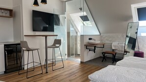 A modern bedroom with a large bed, a desk with two chairs, a TV mounted on the wall, and a bathroom with a mirror and sink.