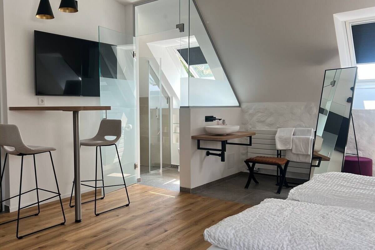 Un dormitorio moderno con una cama grande, un escritorio con dos sillas, un televisor montado en la pared y un baño con espejo y lavabo.